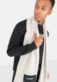 Beige, geribbelde sjaal over een zwarte sweatshirt gedrapeerd. Bevat een zwart label met een wit logo. Het materiaal lijkt zacht en heeft een gladde textuur.