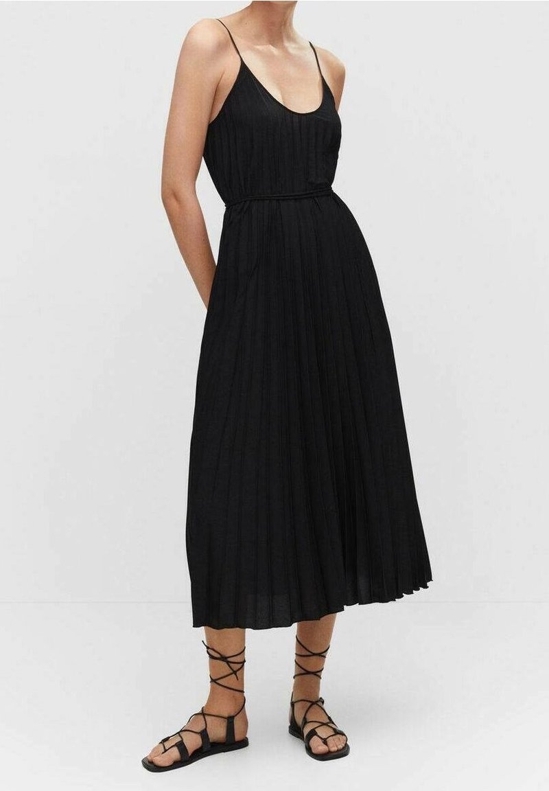 Robe maxi noire plissée avec fines bretelles, taille cintrée et silhouette fluide. Portée avec des sandales plates noires à lanières.