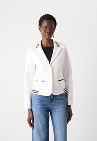 Blazer blanc avec un col cranté, orné de rayures navy, deux poches avant et fermeture à double bouton ; associé à un jean bleu.