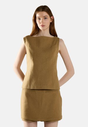 Giovane donna con capelli lunghi che indossa un top marrone senza maniche e una gonna corta abbinata, in piedi davanti a uno sfondo chiaro e uniforme.