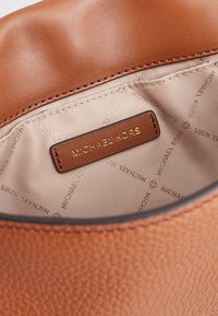 Interno di una borsa in pelle color tan con foderatura beige. Caratterizzata da un'etichetta marrone impressa con "MICHAEL KORS" in oro. Texture liscia e cuciture pulite.