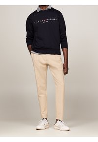 Felpa blu navy con logo bianco "TOMMY HILFIGER", camicia grigia con colletto sotto, pantaloni beige e sneaker bianche. Outfit casual.