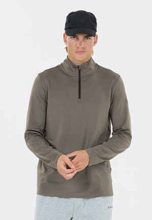 Junger Mann mit schwarzer Kappe, olivgrünem langärmligem Reißverschluss-Shirt und hellgrauer Jogginghose, steht mit vor dem Körper verschränkten Händen.