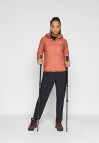 Casaco laranja acolchoado com fecho à frente e capuz, usado sobre uma blusa preta, combinado com calças pretas e botas de caminhada vermelhas, segurando bastões de trekking.