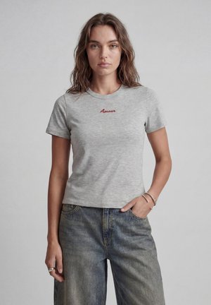 Jeune femme aux cheveux bruns ondulés portant un t-shirt gris clair avec "Amour" en texte rouge et un jean bleu délavé, debout avec une main dans la poche.