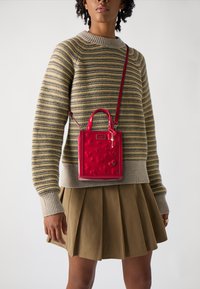 Rode patenten mini-handtas met hartvormige embossing en dubbele handvatten, crossbody gedragen met een lange schouderband tegen een gestreepte trui.