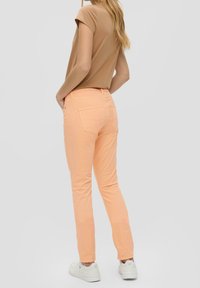 Donna che indossa una maglietta corta marrone chiaro, pantaloni aderenti color pesco e sneakers bianche, in piedi con le mani nelle tasche posteriori.