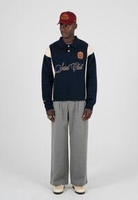 Marineblauer Pullover mit cremefarbenen Akzenten, goldener Stickerei und Polokragen. Graue weit geschnittene Hosen und beige Schuhe. Burgunderfarbene Kappe mit Logodetail.
