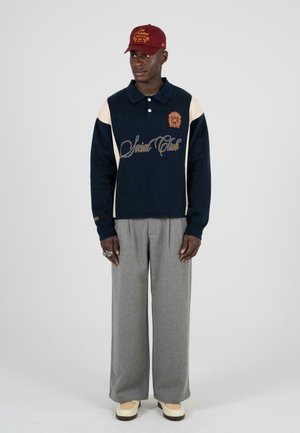 Pull marine avec des accents crème, broderie dorée et col polo. Pantalons gris à larges jambes et chaussures beiges. Casquette bourgogne avec détail de logo.