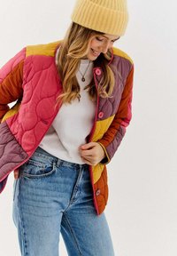 Prešívaná bunda v ružovej, horčicovej a fialovej farbe; má patentné gombíky a mäkký, textúrovaný povrch. Kombinovaná s bielym topom a modrými džínsami.