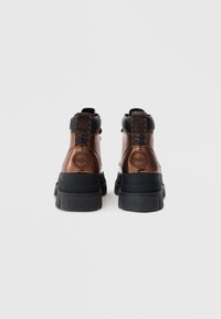 Καφέ ψηλά παπούτσια με γυαλιστερό φινίρισμα, μαύρες λαστιχένιες σόλες και διακριτική λεπτομέρεια λογότυπου στο πίσω λουράκι. Διαθέτουν ραφές και διακοσμητικά στοιχεία.