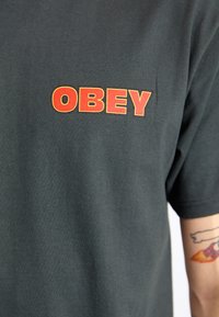 Black cotton T-shirt featuring bold red "OBEY" text on the left chest area. Short sleeves and standard crew neck design. Smooth texture.