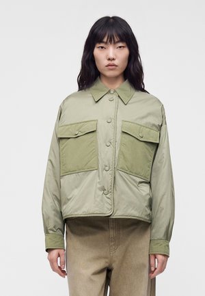 Kvinde med glat mørkt hår iført en lysegrøn knapjakke med store forlommer og beige bukser mod en ensfarvet baggrund.