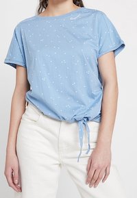 Blå bomullströja med korta ärmar, lös passform och samlad knutdetalj vid kanten. Har små vita blommönster.