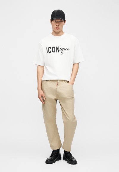 Νεαρός άνδρας που φοράει λευκό T-shirt με την επιγραφή "ICONique", μπεζ παντελόνι, μαύρο καπέλο και μαύρα αρβυλάκια με κορδόνια, στέκεται μπροστά σε απλό φόντο.