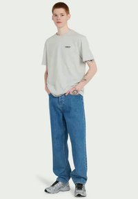 Pelēks kokvilnas T-krekls ar īsām piedurknēm un krūšu logotipu, apvienots ar brīviem zilas džinsu biksēm un sudrabainām sporta kurpēm.