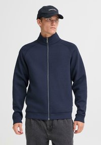 Navy zip-up sweatshirt with a high collar, long sleeves, textured fabric, and contrasting zipper. Paired with dark gray pants and a cap.