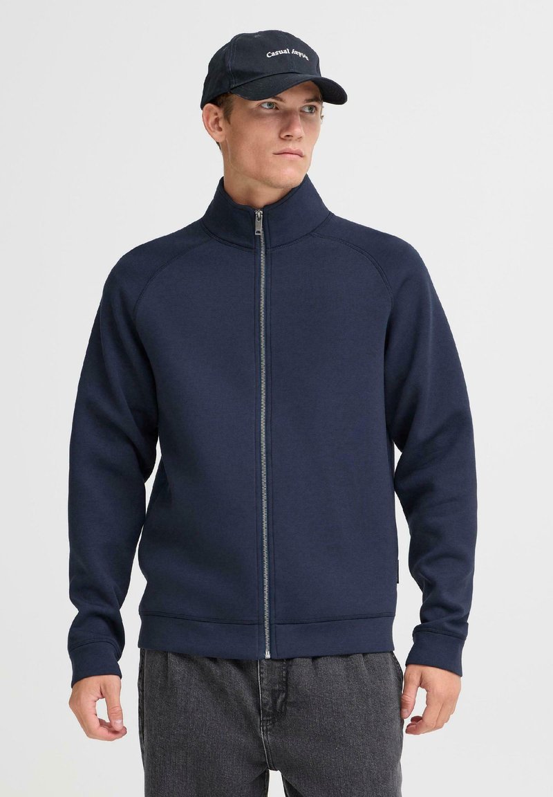 Felpa zip-up blu navale con colletto alto, maniche lunghe, tessuto strutturato e zip a contrasto. Abbinata a pantaloni grigi scuri e a un cappellino.