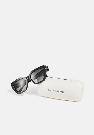 Occhiali da sole neri con montatura angolare e lenti grigie, posizionati accanto a un astuccio bianco impresso con "Alexander McQUEEN." Design elegante e superfici testurizzate.