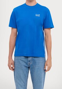 Modré bavlněné tričko s krátkými rukávy, kulatým výstřihem, bílým logem na hrudi a bílými akcentními pruhy po stranách. Nosit se světle modrými džíny.