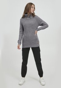 Grauer Strickpullover mit hohem Kragen, geripptem Textur und Seitentaschen, kombiniert mit schwarzen Cargo-Hosen und weißen Turnschuhen.