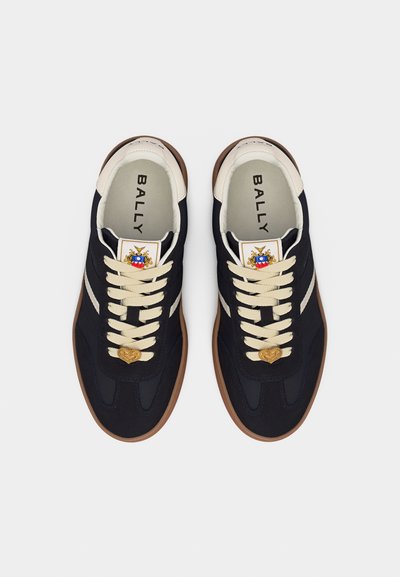 Paire de baskets noires Bally avec lacets crème et col de talon, semelles marron, et un pendentif en forme de cœur sur les lacets, vue de dessus.