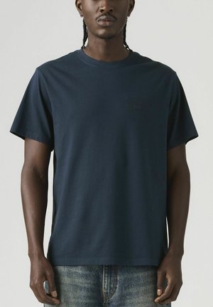 Mężczyzna z zaplecionymi włosami, ubrany w prostą ciemnogranatową koszulkę z krótkim rękawem i niebieskie dżinsy, stojący na tle o neutralnym kolorze.