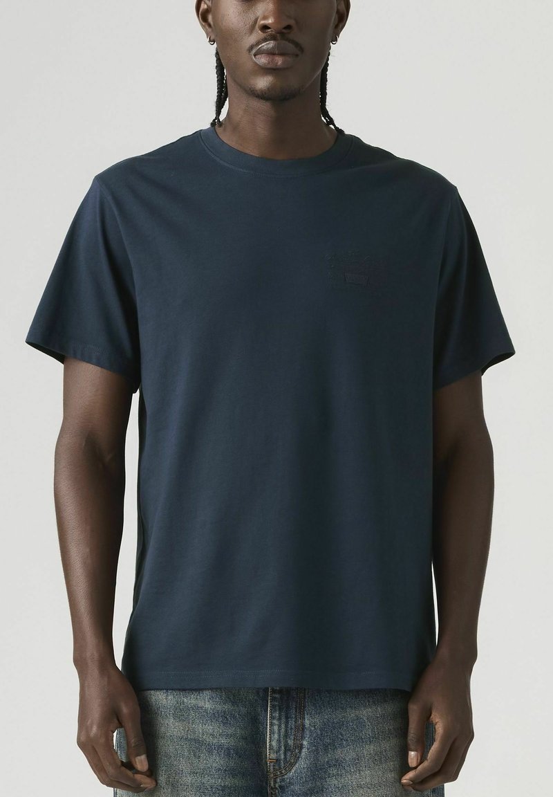 Hombre con cabello trenzado que lleva una camiseta de manga corta azul marino lisa y jeans azules, de pie frente a un fondo neutro.