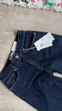 Σκούρο μπλε τζιν με μπροστινές τσέπες και προσαρτημένες λευκές ετικέτες τοποθετημένα σε ξύλινο δάπεδο κοντά στην άκρη υφασμάτινου χαλιού.