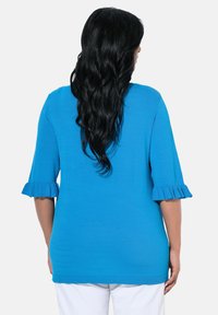 Blaues Oberteil mit Dreiviertelärmeln und Rüschendetails, aus strukturiertem Stoff und geradem Schnitt. Getragen mit weißen Hosen.