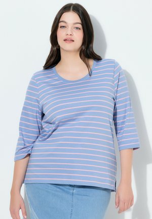 Femme aux longs cheveux foncés portant un chemisier à manches 3/4 bleu et rose à rayures et une jupe en denim bleu clair, sur fond blanc uni.