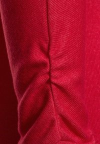 Roter Strickstoff mit einem strukturierten Muster und Raffungen an den Ellbogen, der das glatte und weiche Material hervorhebt.
