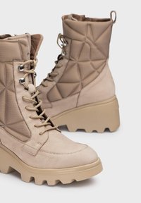 Botas de tobillo beige hechas de cuero y textil con un diseño acolchado, parte delantera con cordones y suela gruesa y texturizada.