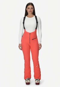 Pantalon de ski corail avec bretelles noires réglables, fermeture éclair à l'avant et poche latérale. Coupe ajustée avec texture adaptée à la neige. Haut blanc à manches longues visible.