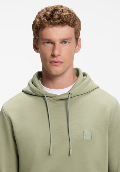 Jeune homme aux cheveux courts et bouclés portant un sweat à capuche vert clair avec des cordons et un petit logo carré sur la poitrine.