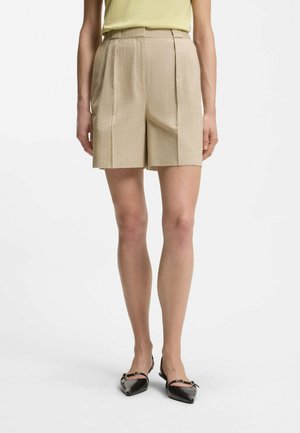Frau trägt beige Nadelstreifen-Hochbundhose, gelbes Oberteil und schwarze spitze Slingback-Ballerinas, steht vor weißem Hintergrund.