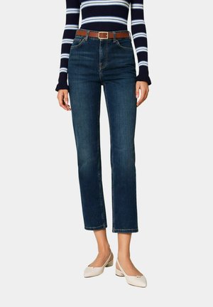 Donna che indossa jeans blu scuro a gamba dritta con cintura marrone, top a maniche lunghe a righe blu navy e bianco e scarpe bianche con tacco basso.