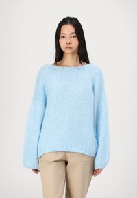 Lichtblauwe oversized gebreide trui met een brede halslijn, lange mouwen en een gestructureerde afwerking. Gepaard met beige broeken.