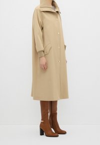 Abrigo beige de gran tamaño con un alto cuello, grandes botones y bolsillos laterales; combinado con botas de cuero marrón hasta la rodilla con tacón bloque.