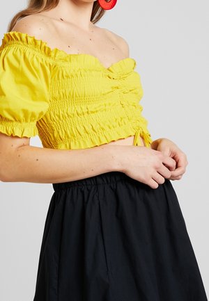 Frau trägt ein gelbes, schulterfreies, gerafftes Crop-Top mit Puffärmeln und einen schwarzen, hoch taillierten Rock und hält ihre Hände in der Nähe ihrer Taille.