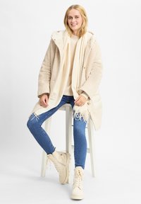 Cappotto di pile beige con cappuccio, sciarpa crema, maglione crema, jeans blu e stivali alti beige con dettagli in cerniera.