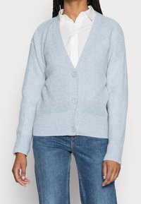 Ljusblå stickad cardigan med v-ringad hals, prydd med fem knappar och ribbade ärmslut, kombinerad med en vit skjorta och denimjeans.