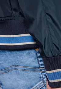 Granatowa kurtka przeciwwiatrowa z prążkowanym mankietem w poziome niebieskie i kremowe paski, w połączeniu z jasnoniebieskimi dżinsami. Gładka tekstura i luźny krój.