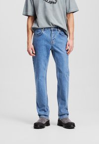 Jean en denim bleu clair à coupe droite, cinq poches et détails de couture ; porté avec une chemise grise et des bottes grises.