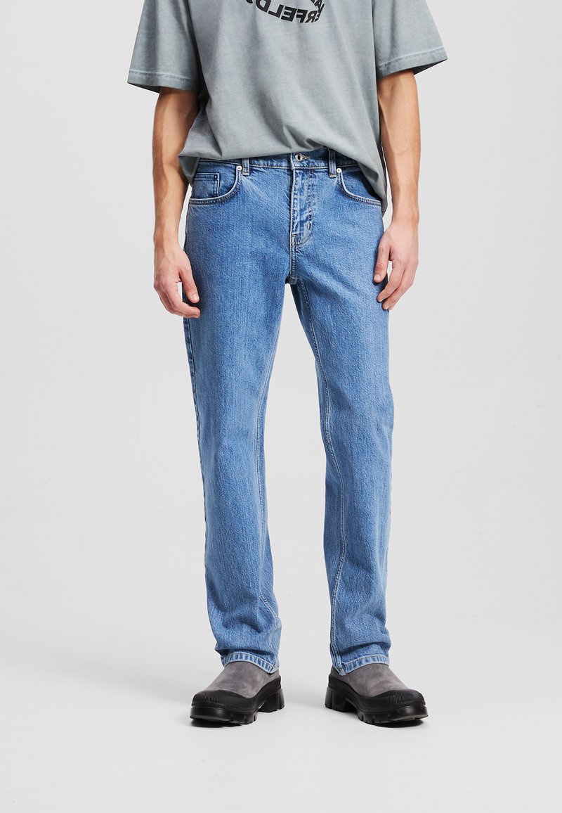 Jean en denim bleu clair à coupe droite, cinq poches et détails de couture ; porté avec une chemise grise et des bottes grises.