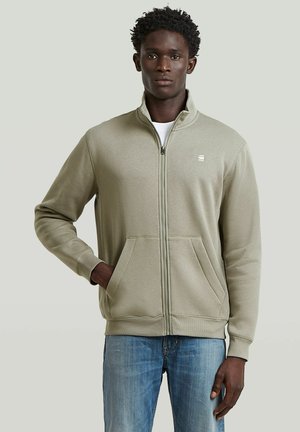 Jonge man die een lichtbeige jas met rits en zakken en een blauwe spijkerbroek draagt, staand tegen een effen lichtgrijze achtergrond.