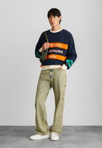 Sweatshirt bleu marine avec des rayures orange et blanche ; jeans en denim clair à coupe décontractée ; baskets blanches ; sac bandoulière vert olive ; poignets texturés.