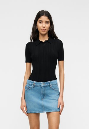 Jauna moteris su ilgais tamsiais plaukais dėvi juodą trumpomis rankovėmis megztą siūlių viršutinę dalį ir šviesiai mėlyną džinsinę mini sijoną, stovinti priešais paprastą foną.