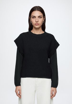 Femme aux cheveux lisses et foncés portant des boucles d'oreilles créoles dorées, un haut en maille noir sans manches sur une chemise noire à manches longues et un pantalon blanc, regardant vers l'avant.