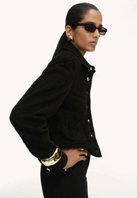 Giacca in pelle scamosciata nera con bottoni a pressione, tasche laterali e colletto strutturato. Indossata con jeans neri e bracciale in oro. Fit slim e su misura.